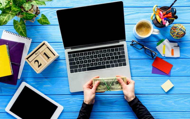 hands holding money by laptop on blue wooden table with stationery